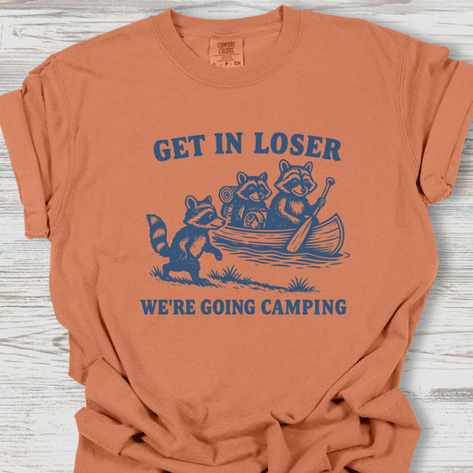 Orange t-shirt with blue graphic and text 'Get in Loser, We're Going Camping' on a wooden surface