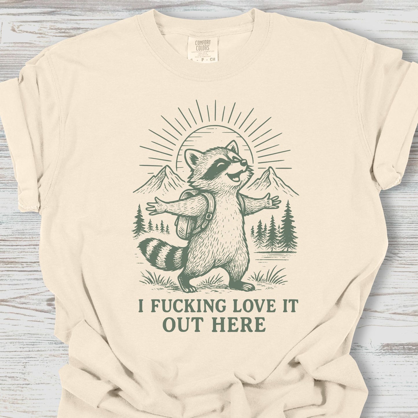 Beige t-shirt with a raccoon graphic and text on a wooden surface