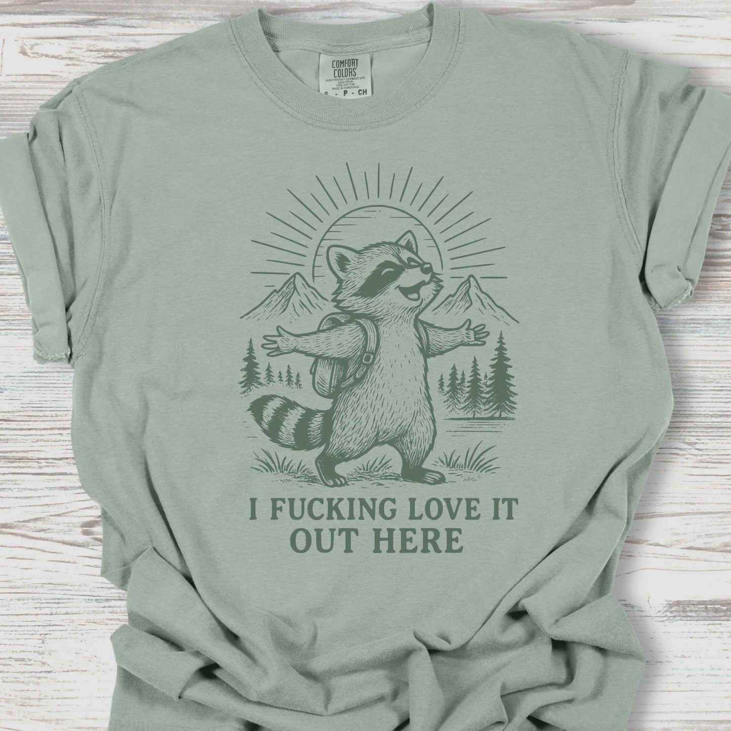 Sage green t-shirt with a raccoon graphic and text on a wooden surface