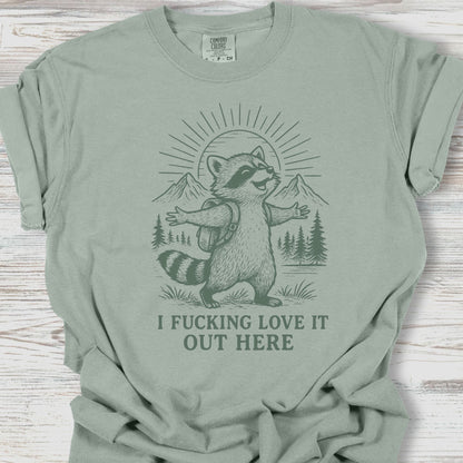 Sage green t-shirt with a raccoon graphic and text on a wooden surface