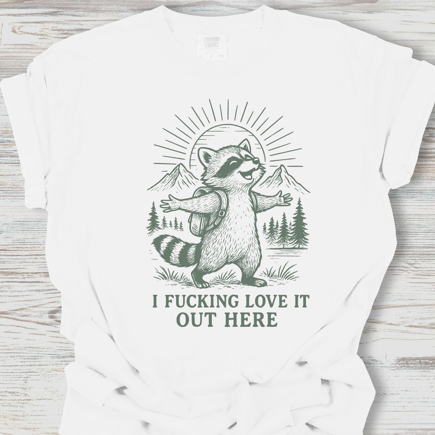 White t-shirt with a raccoon graphic and text on a wooden surface