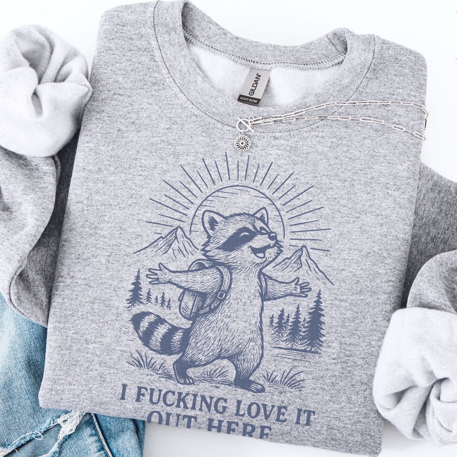 Gray sweatshirt with a raccoon graphic and text, displayed with jeans on a white background.