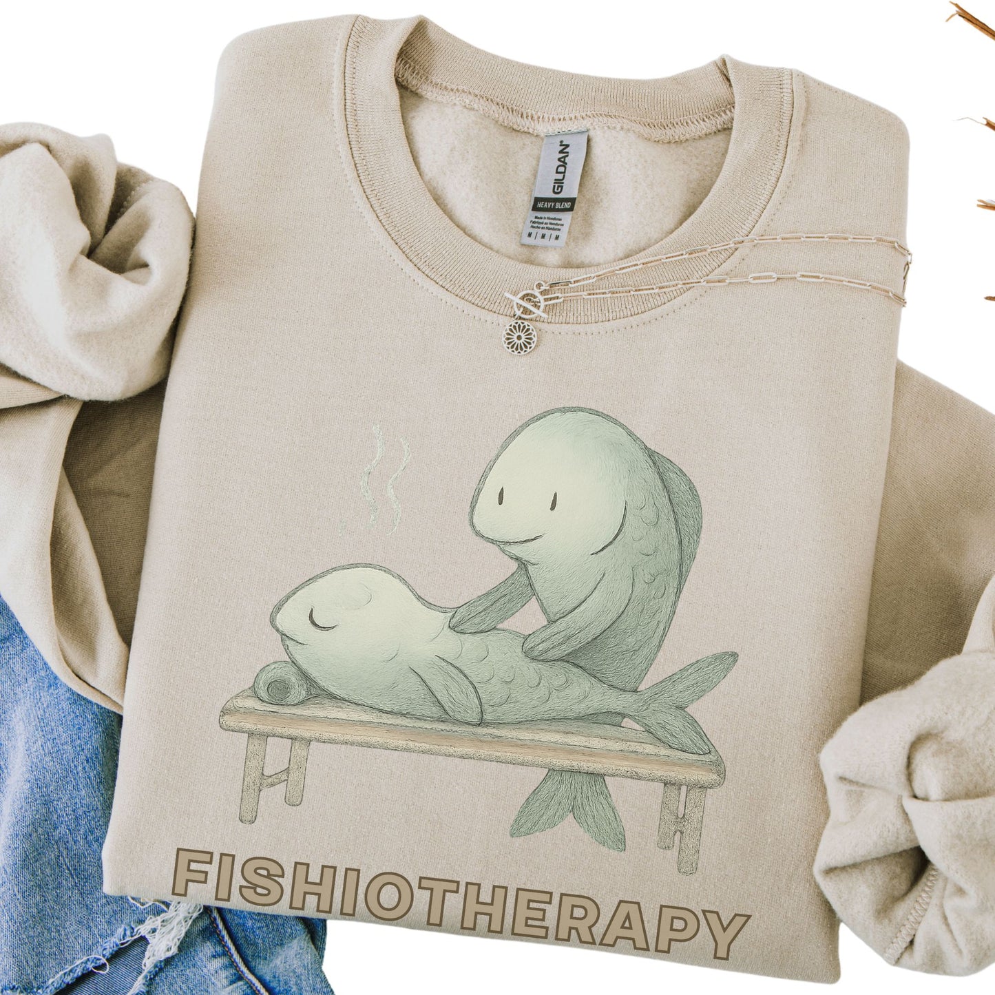 Beige sweatshirt with 'Fishiotherapy' design featuring two fish on a bench, held by a person.