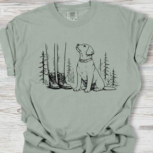 Sage green t-shirt with a graphic of a dog and boots in a forest on a wooden surface.