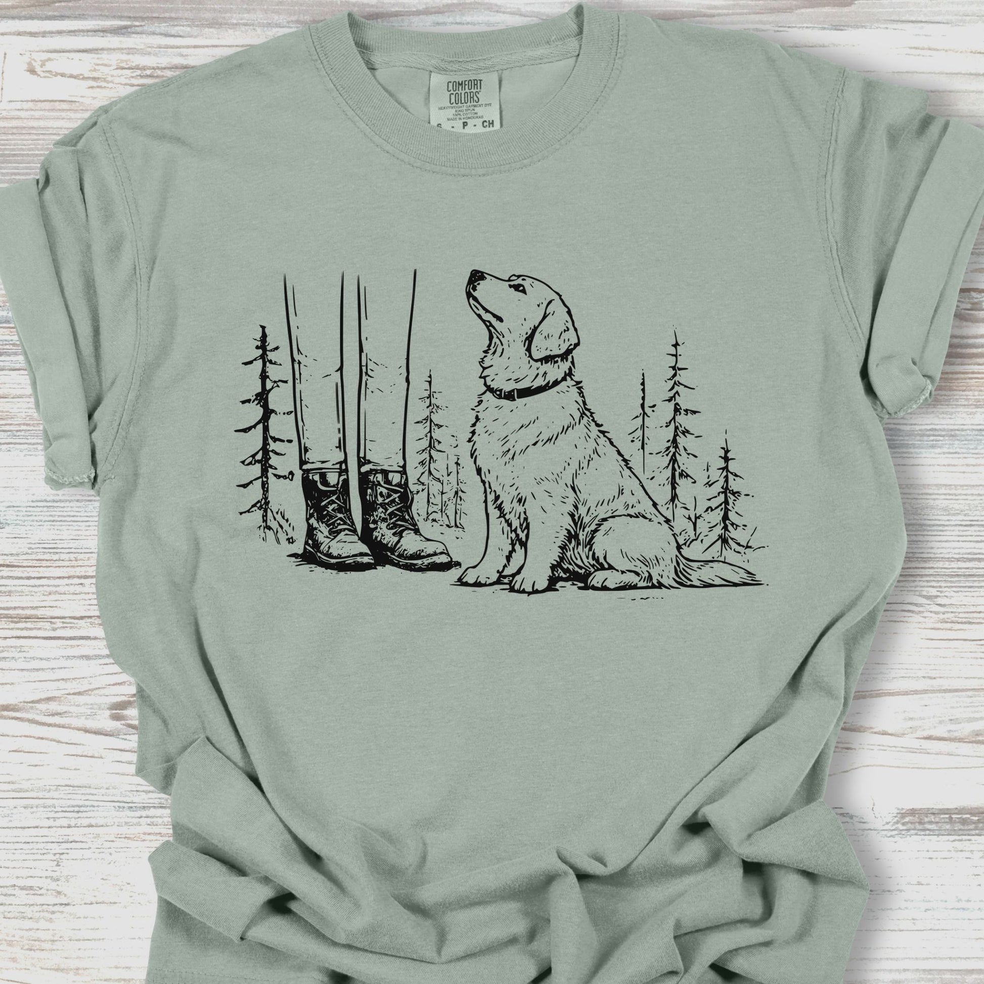 Sage green t-shirt with a graphic of a dog, boots, and trees on a wooden surface