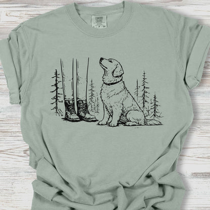 Sage green t-shirt with a graphic of a dog, boots, and trees on a wooden surface