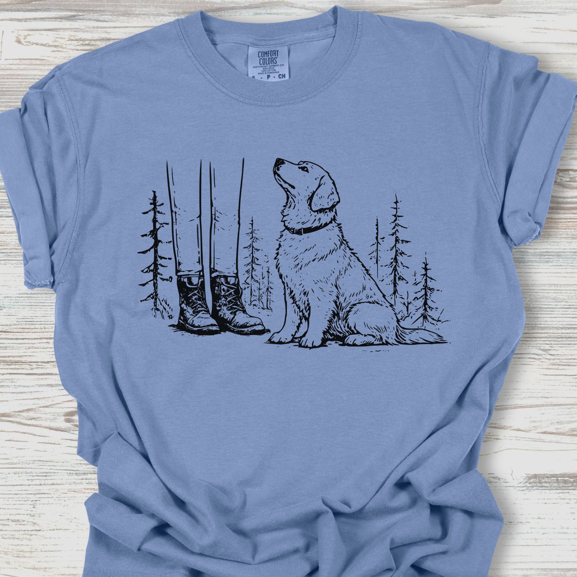 Blue t-shirt with a graphic of a dog, boots, and trees on a wooden surface