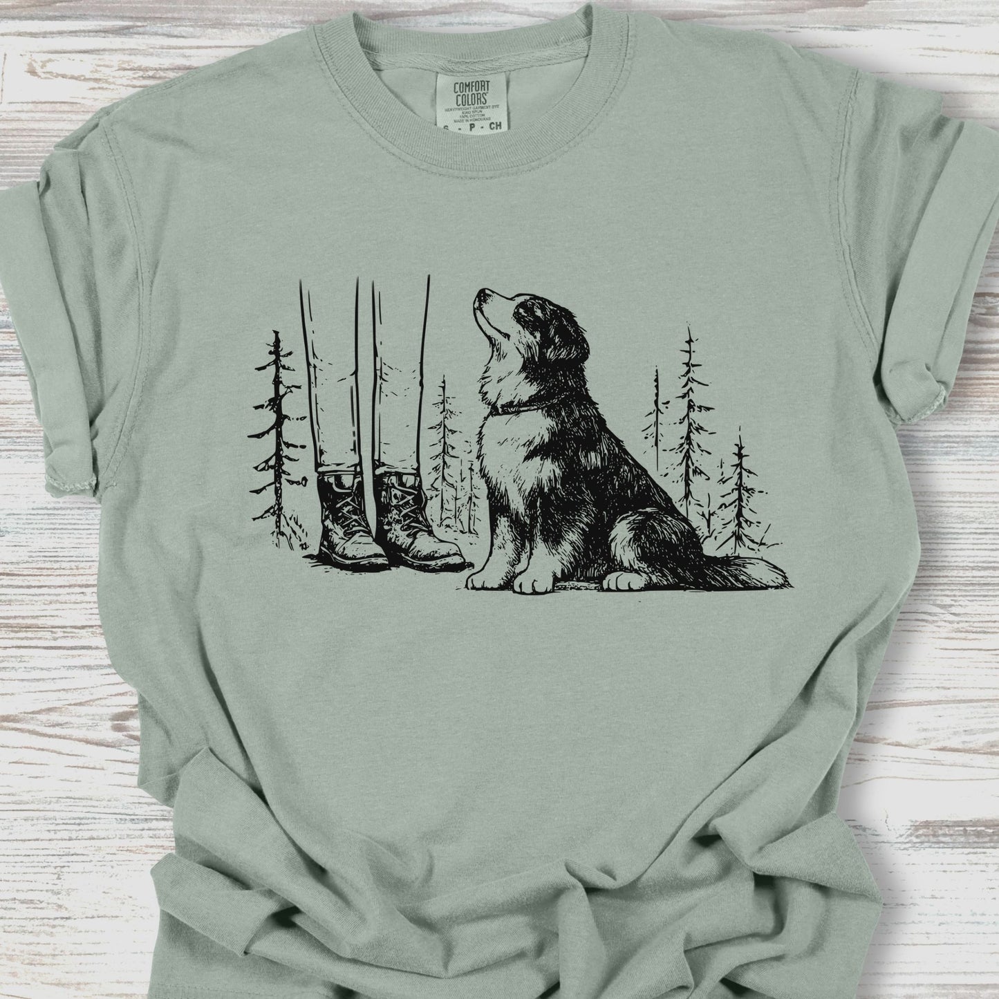 Sage green t-shirt with a graphic of a dog, boots, and trees on a wooden surface