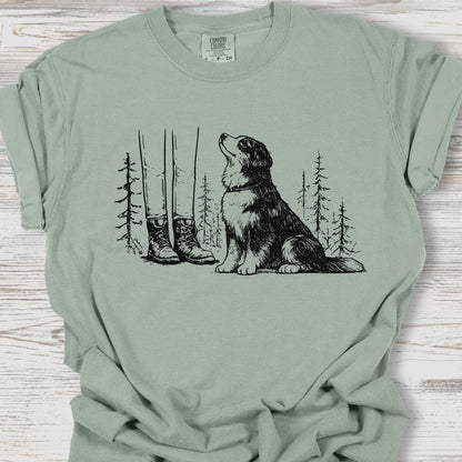 Sage green t-shirt with a graphic of a dog, boots, and trees on a wooden surface