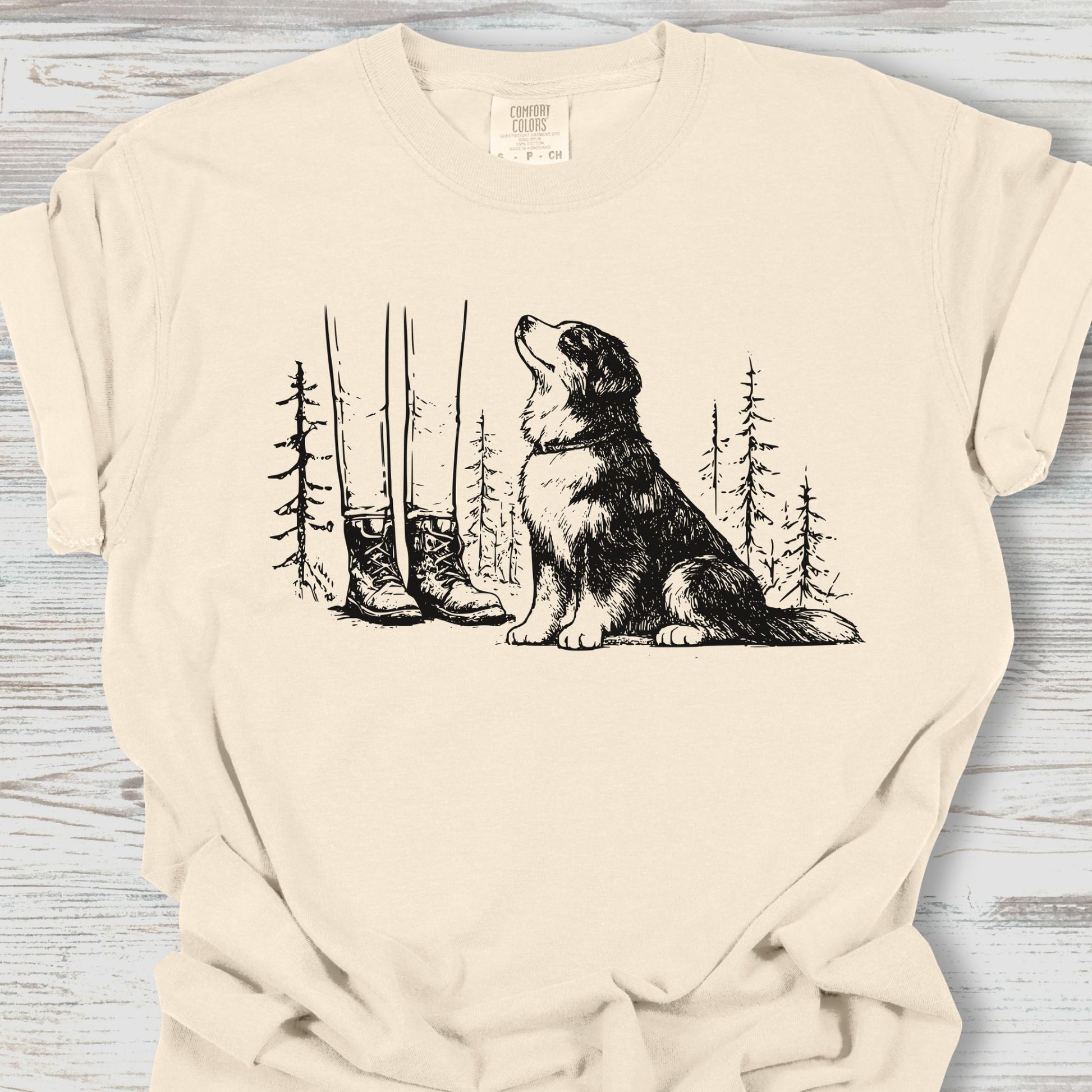 Beige t-shirt with a graphic of a dog and boots in a forest on a wooden surface