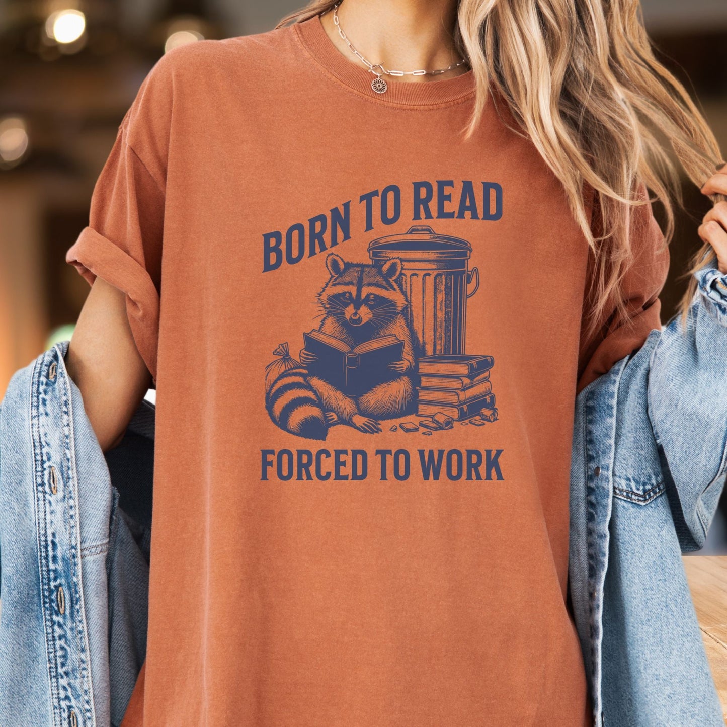 Model wears a Yam colored tshirt with the image of a racoon reading a book in front of a garbage can.  The lettering reads Born to Read, Forced to Work.