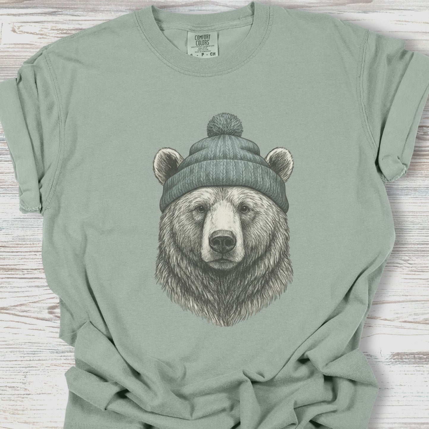 Sage green t-shirt with a bear wearing a beanie graphic on a wooden surface