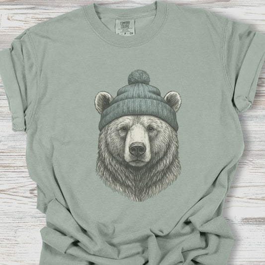 Sage green t-shirt with a bear wearing a beanie graphic on a wooden surface