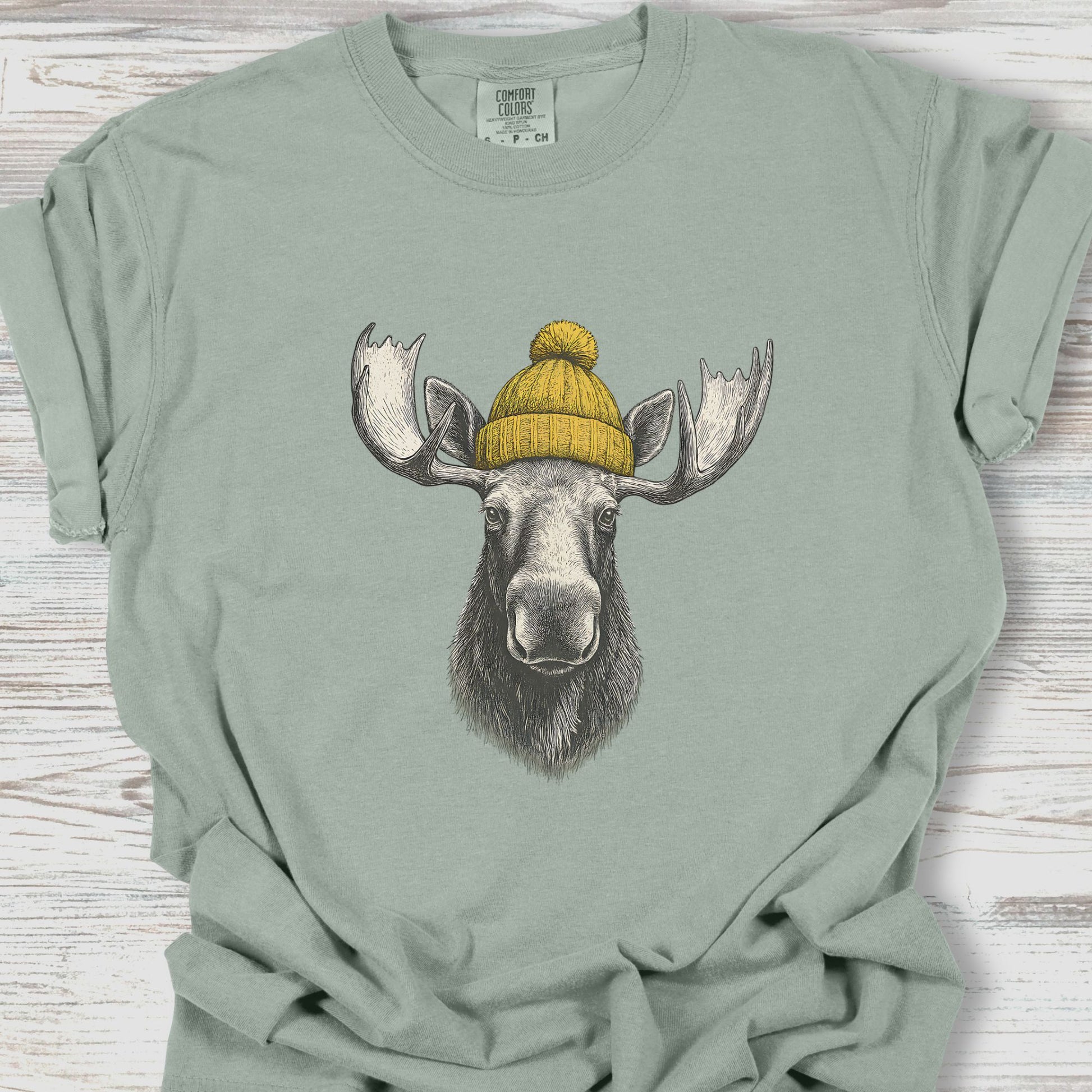 Green t-shirt with a moose wearing a yellow beanie on a wooden surface