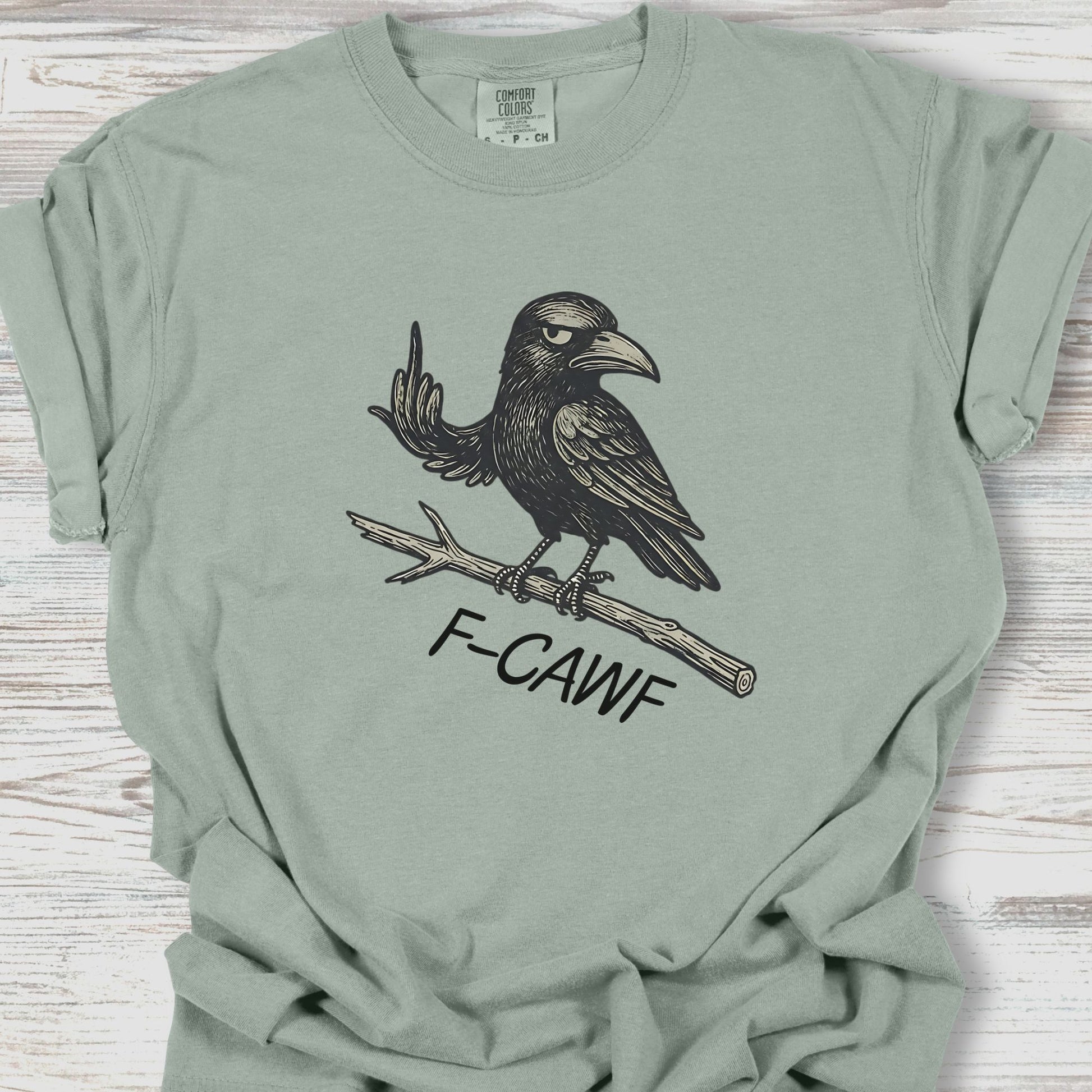 Sage green t-shirt with a crow graphic and 'F-CAWF' text on a wooden surface