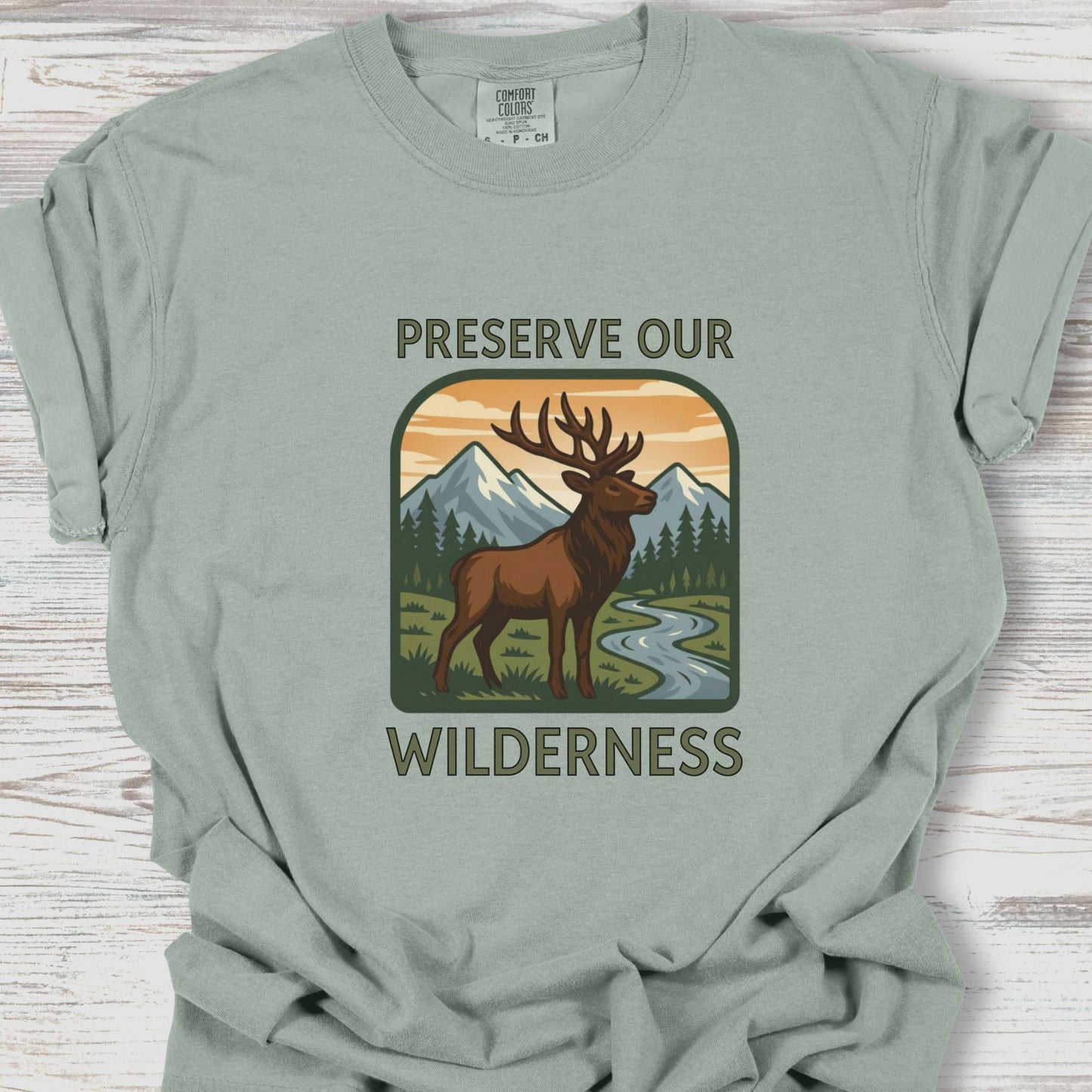 Sage green t-shirt with a graphic of a deer and mountains and text 'Preserve Our Wilderness' on a wooden surface.