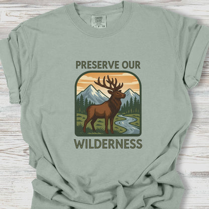 Sage green t-shirt with a graphic of a deer and mountains and text 'Preserve Our Wilderness' on a wooden surface.