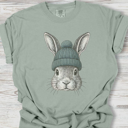 Sage green t-shirt with a graphic of a rabbit wearing a beanie on a wooden surface