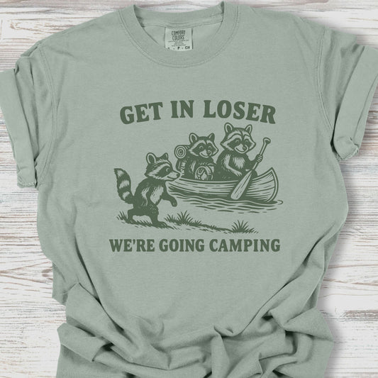 Sage green t-shirt with raccoon graphic and text 'Get in loser, we're going camping' on a wooden surface.