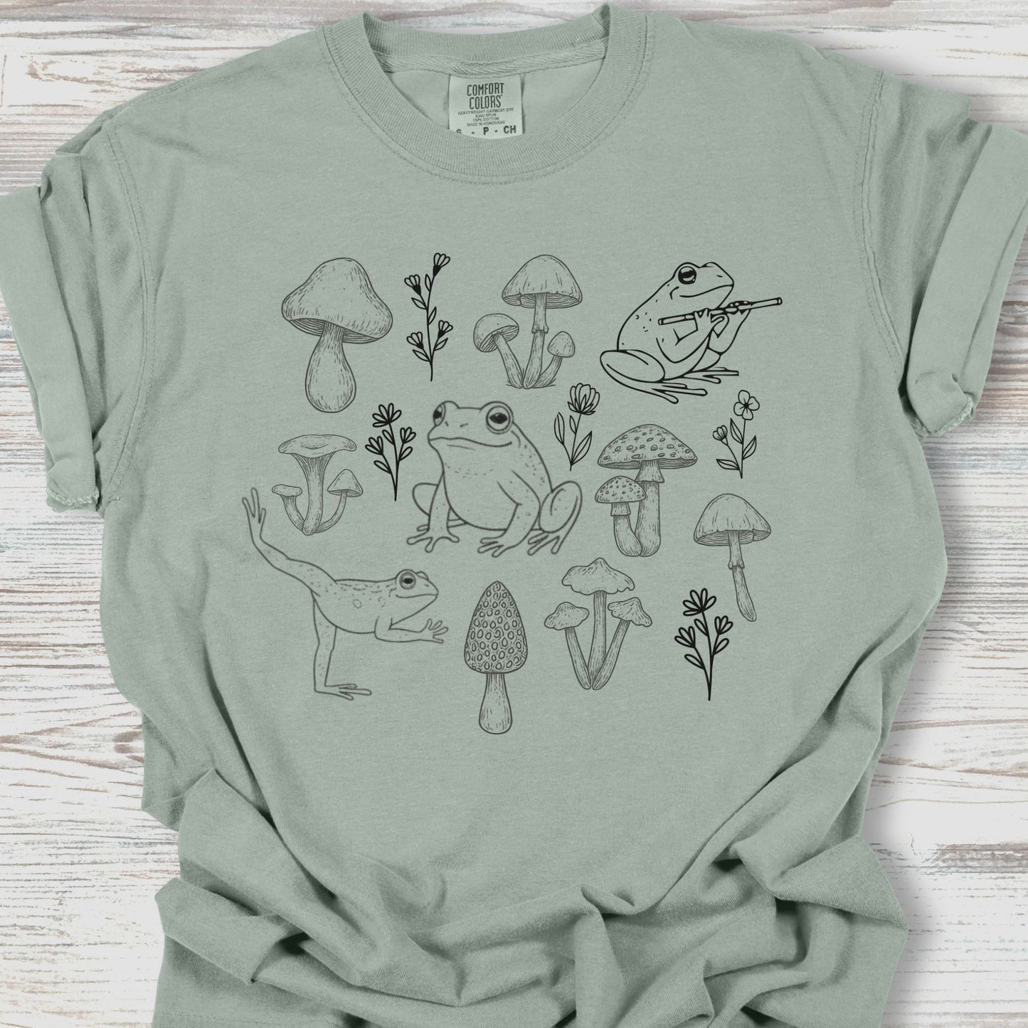 Sage green t-shirt with black line art of frogs and mushrooms on a wooden surface