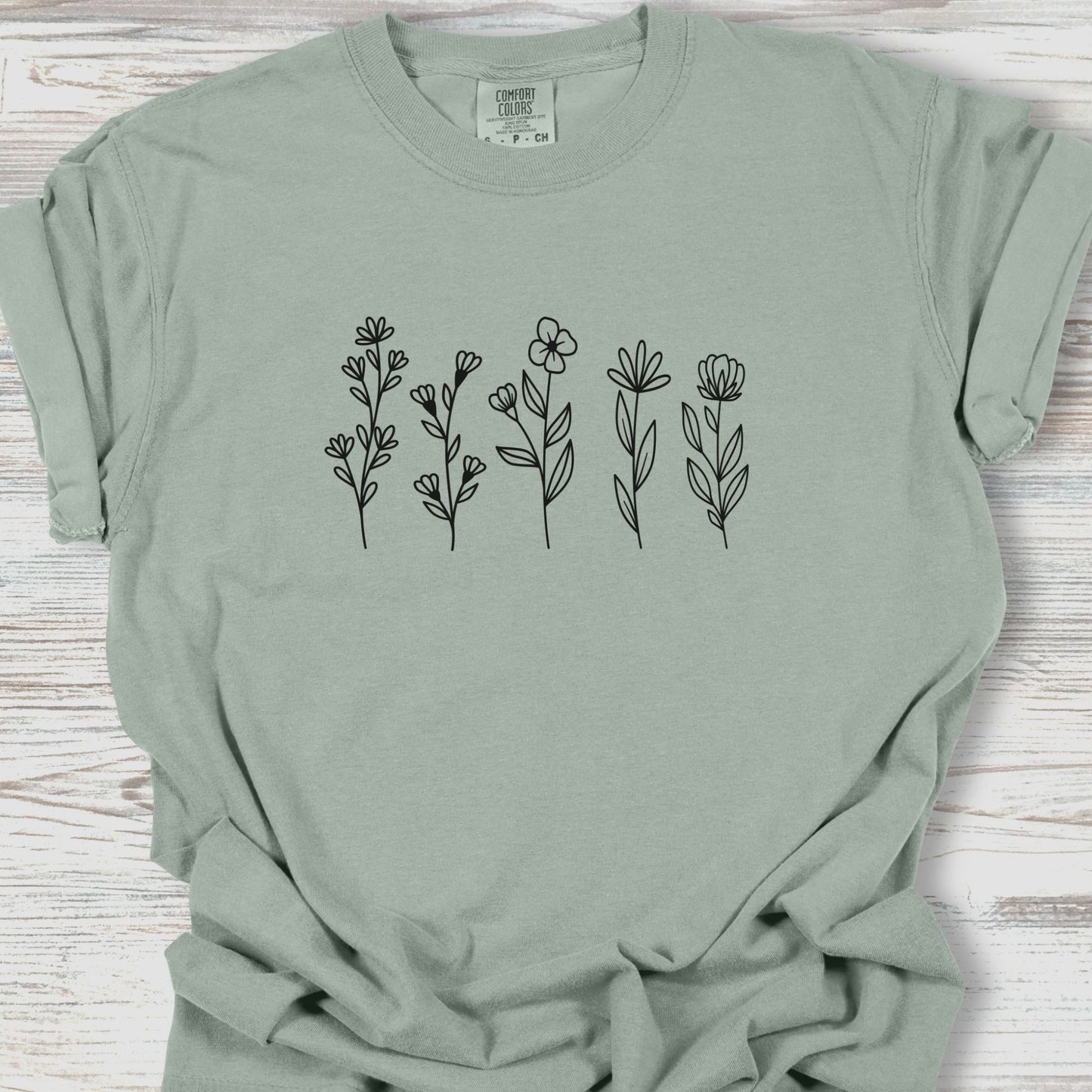 Sage green t-shirt with black floral designs on a wooden surface