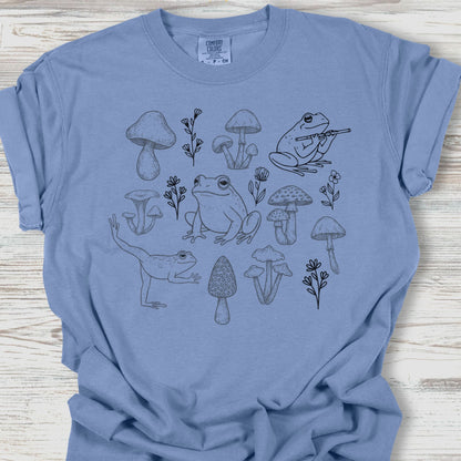 Blue t-shirt with black frog and mushroom design on a wooden surface