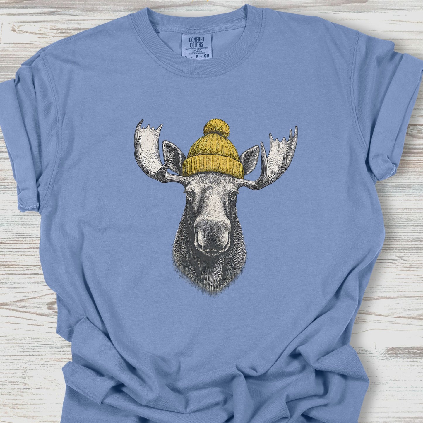Blue t-shirt with a moose wearing a yellow beanie on a wooden surface