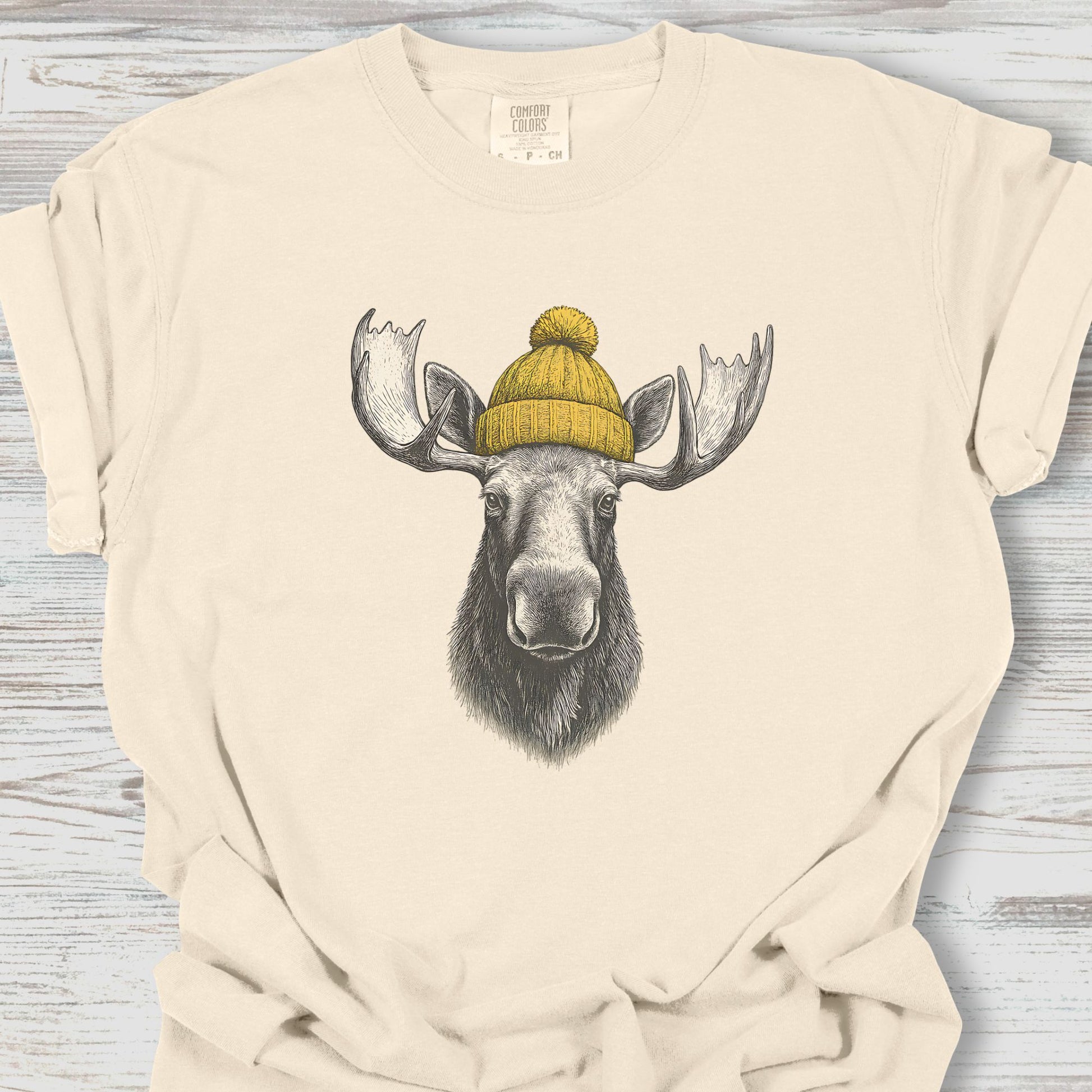 Beige t-shirt with a graphic of a moose wearing a yellow beanie on a wooden surface