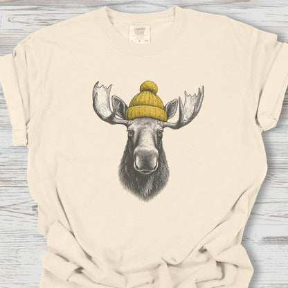 Beige t-shirt with a graphic of a moose wearing a yellow beanie on a wooden surface