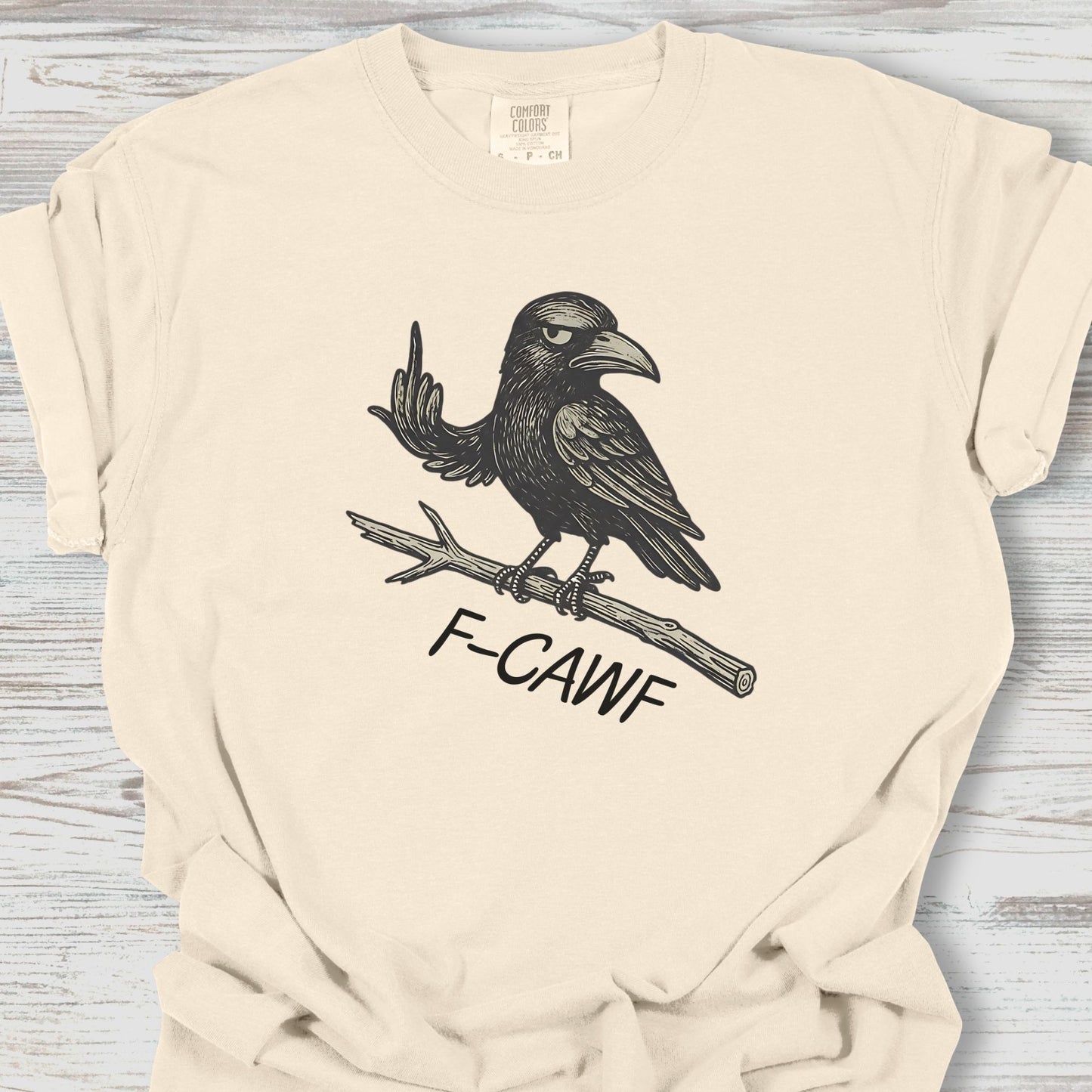 Beige t-shirt with a graphic of a crow and 'F-CAWF' text on a wooden surface