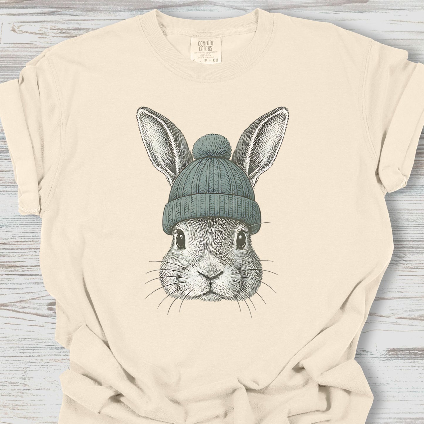 Beige t-shirt with a graphic of a rabbit wearing a blue knit hat on a wooden surface.