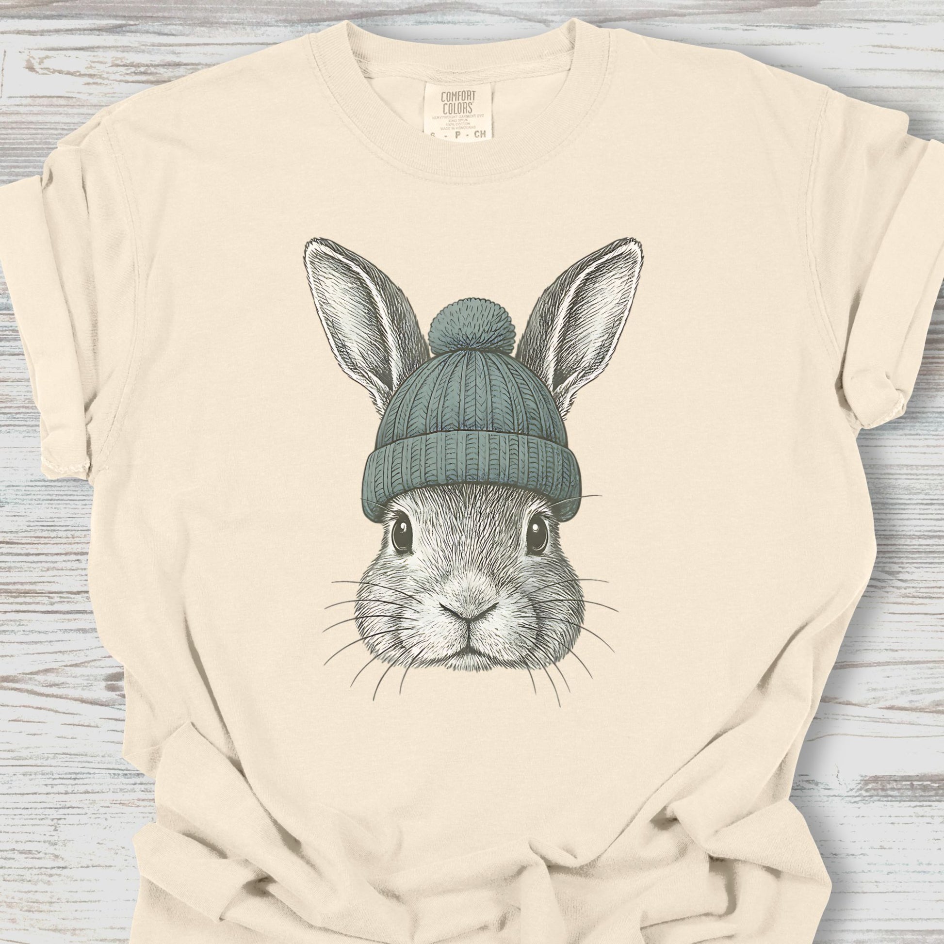 Beige t-shirt with a graphic of a rabbit wearing a blue knit hat on a wooden surface.