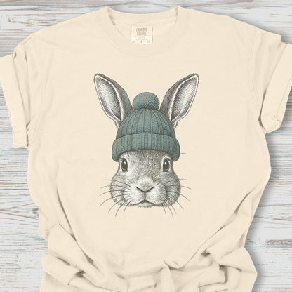 Beige t-shirt with a graphic of a rabbit wearing a blue knit hat on a wooden surface.