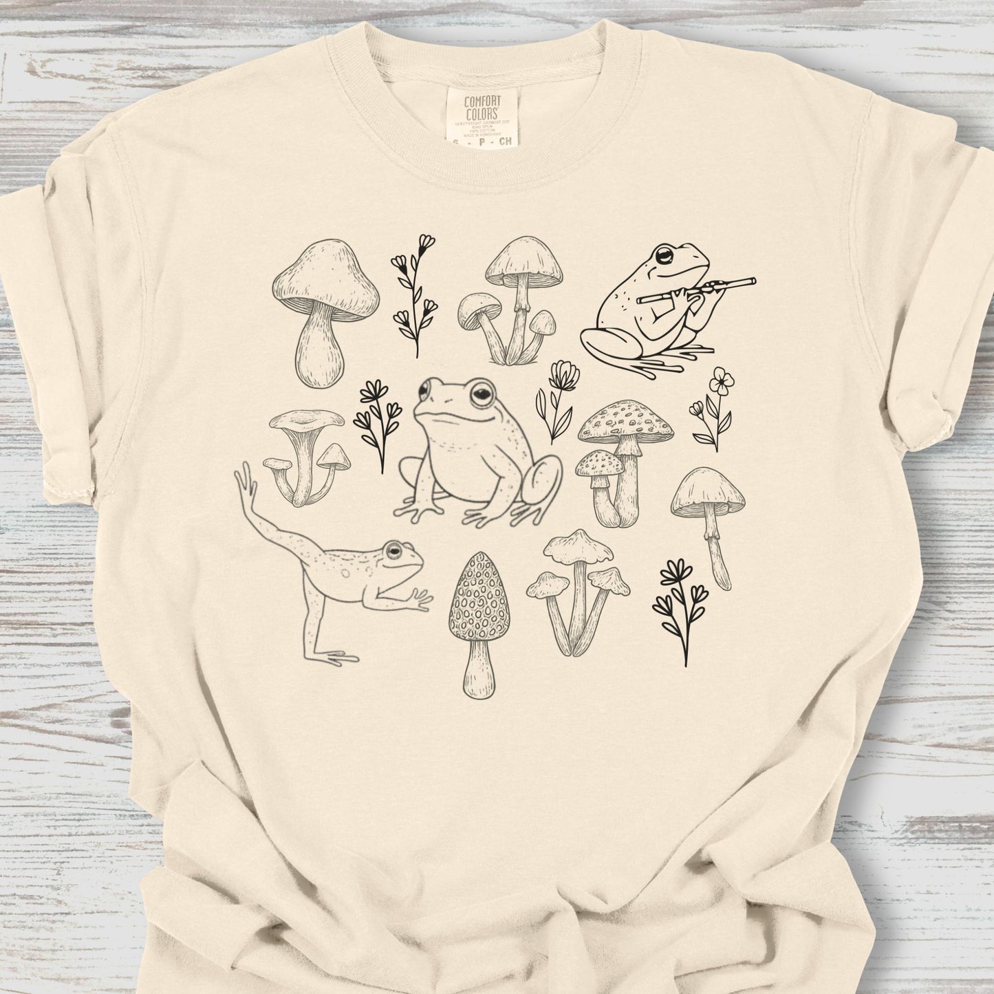 Beige t-shirt with black line art of frogs, mushrooms, and plants on a wooden surface