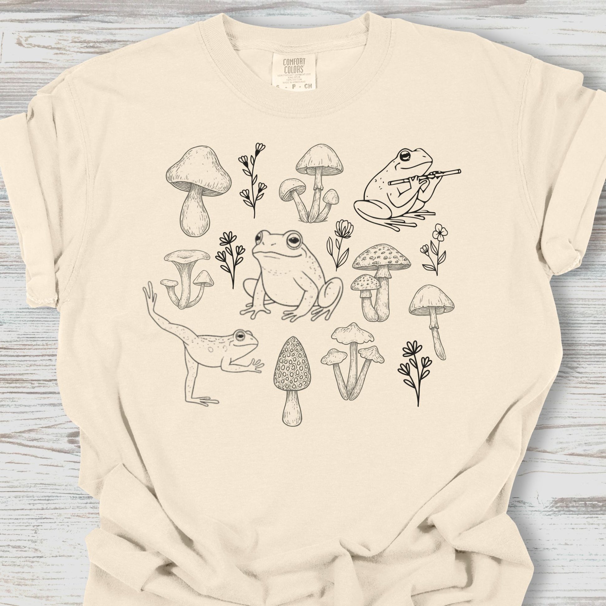 Beige t-shirt with black line art of frogs, mushrooms, and plants on a wooden surface