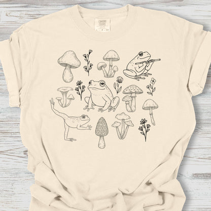 Beige t-shirt with black line art of frogs, mushrooms, and plants on a wooden surface