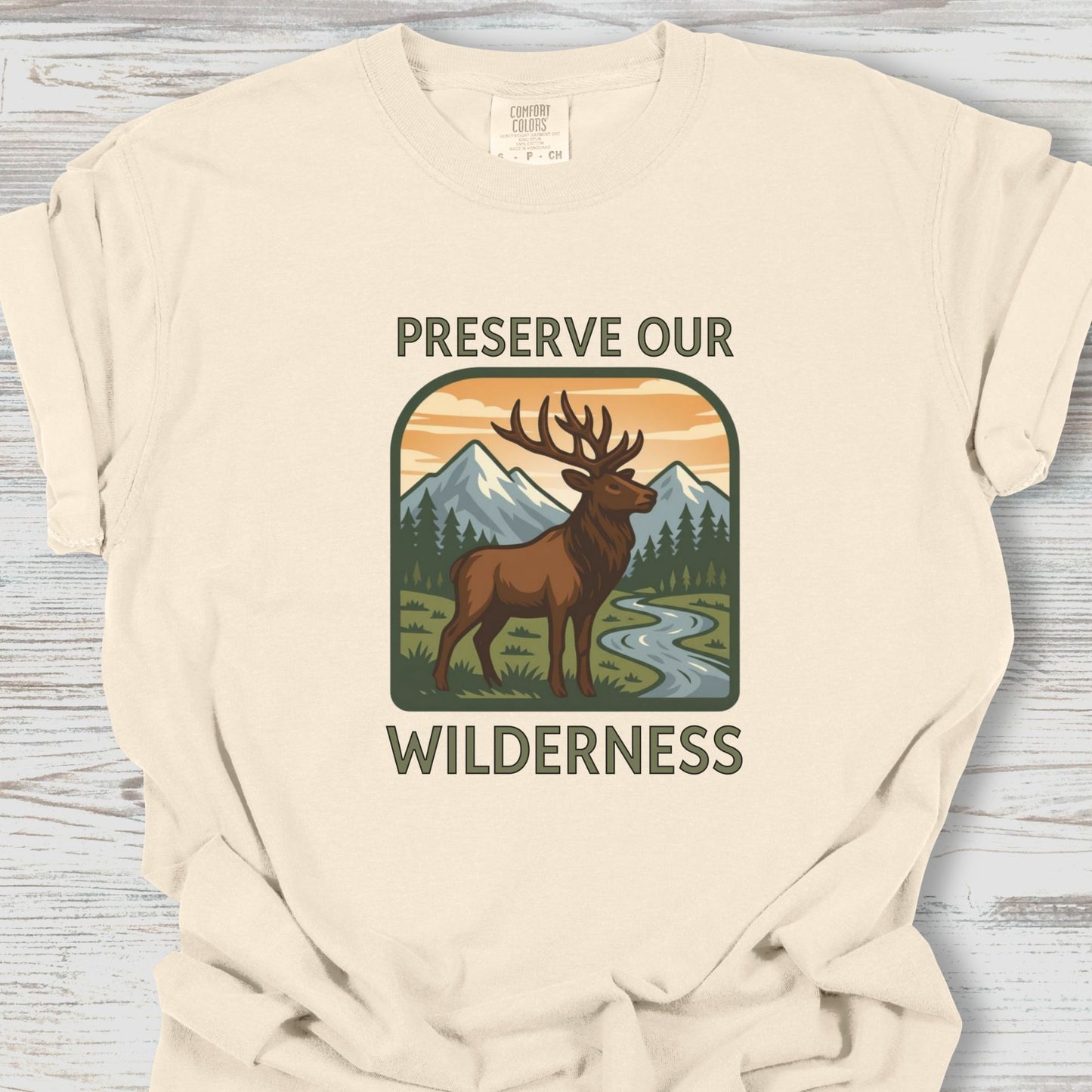 Beige t-shirt with a graphic of a deer and mountains and text 'Preserve Our Wilderness' on a wooden surface.