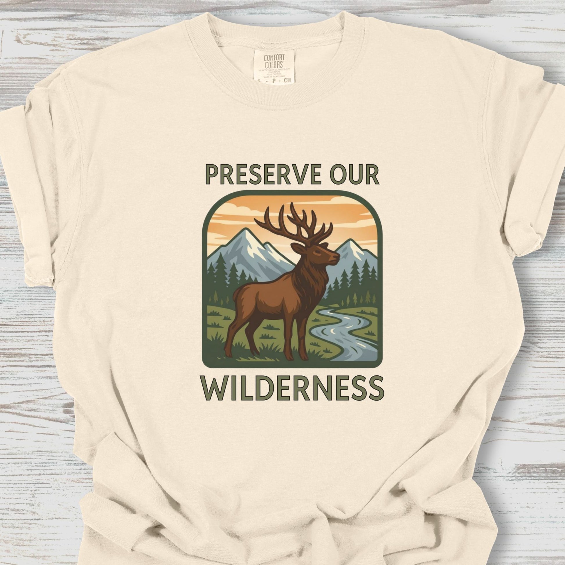 Beige t-shirt with a graphic of a deer and mountains and text 'Preserve Our Wilderness' on a wooden surface.