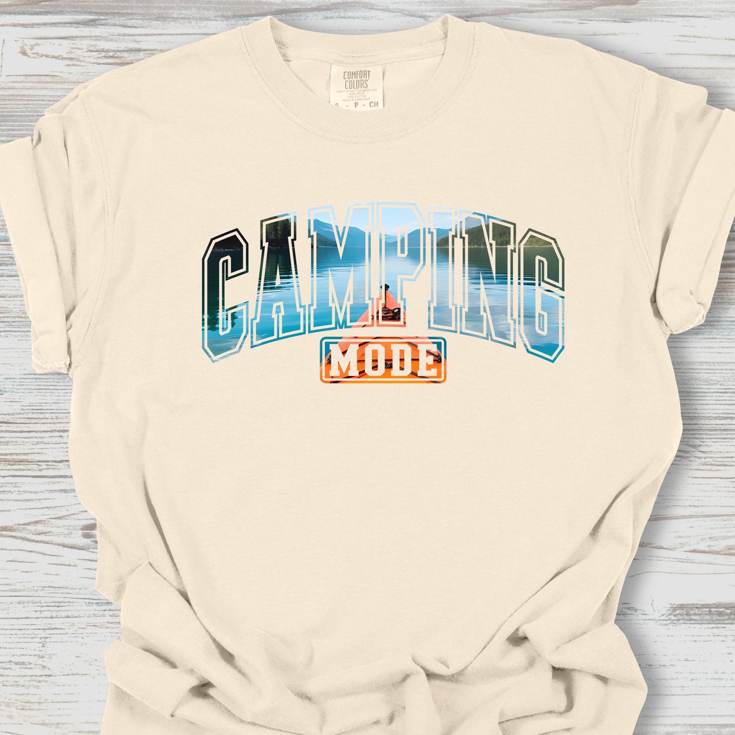 Beige t-shirt with 'Camping Mode' graphic on a wooden surface