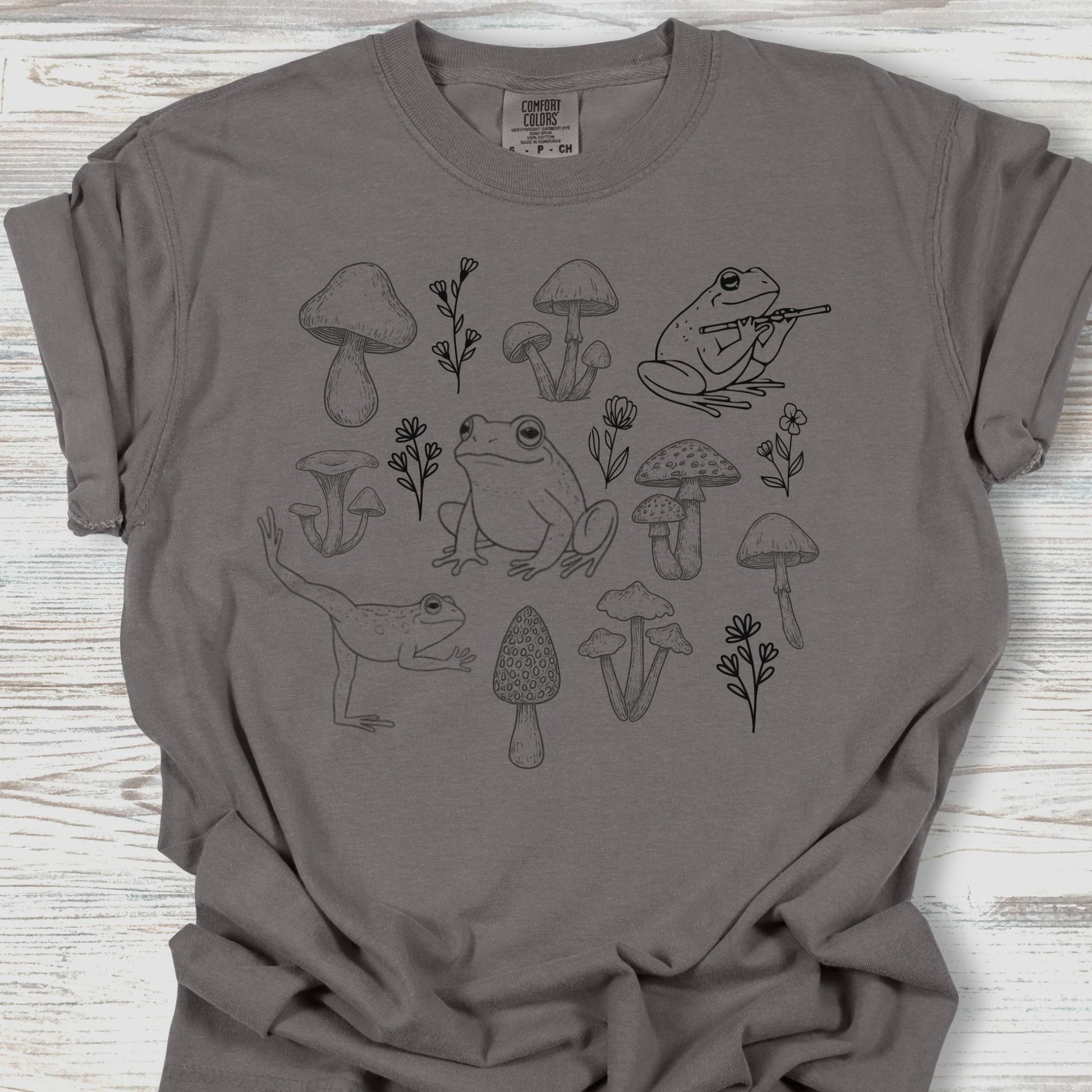 Gray t-shirt with black frog and mushroom design on a wooden surface