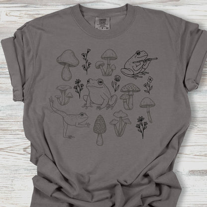 Gray t-shirt with black frog and mushroom design on a wooden surface