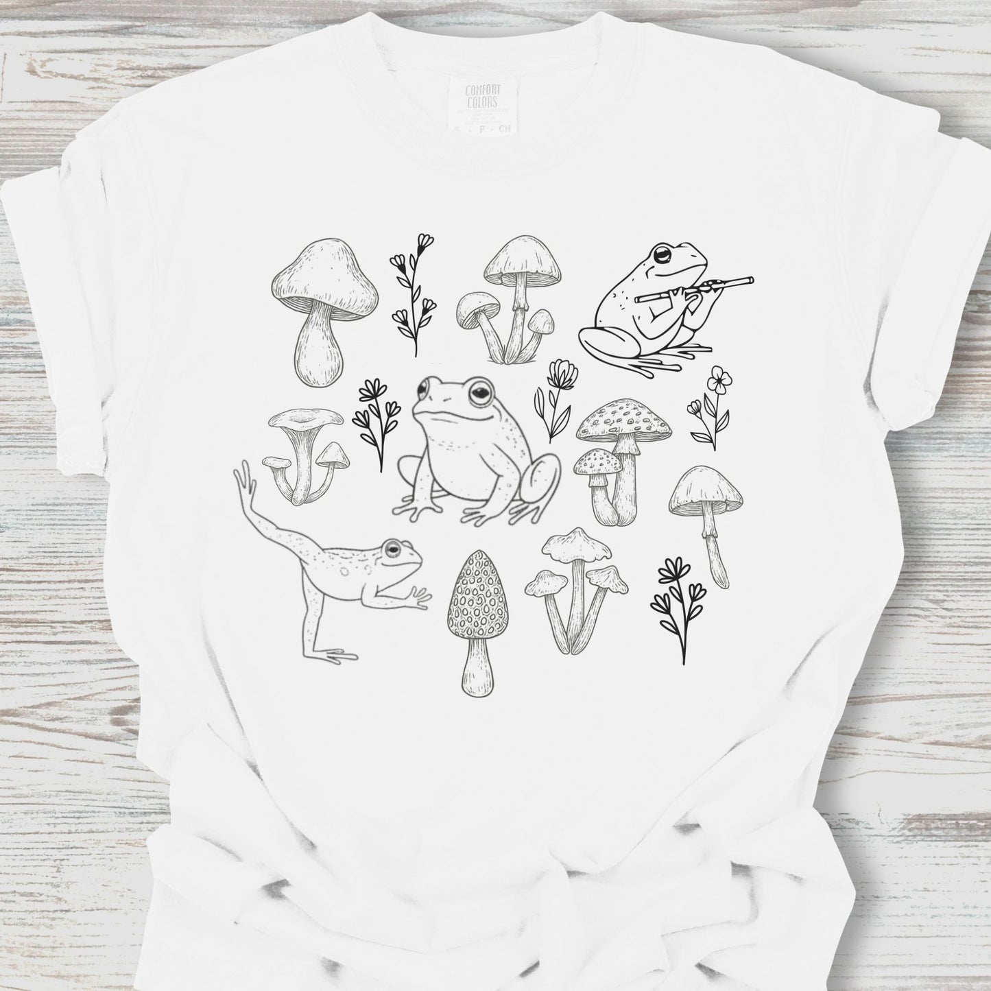 White t-shirt with black line art design of frogs and mushrooms on a wooden surface