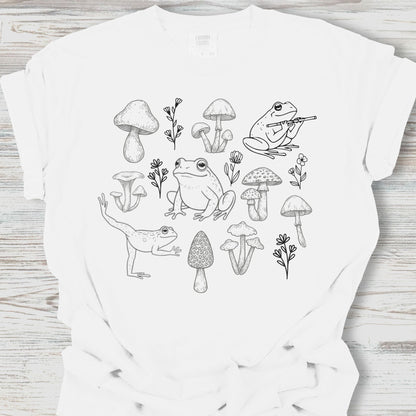 White t-shirt with black line art design of frogs and mushrooms on a wooden surface
