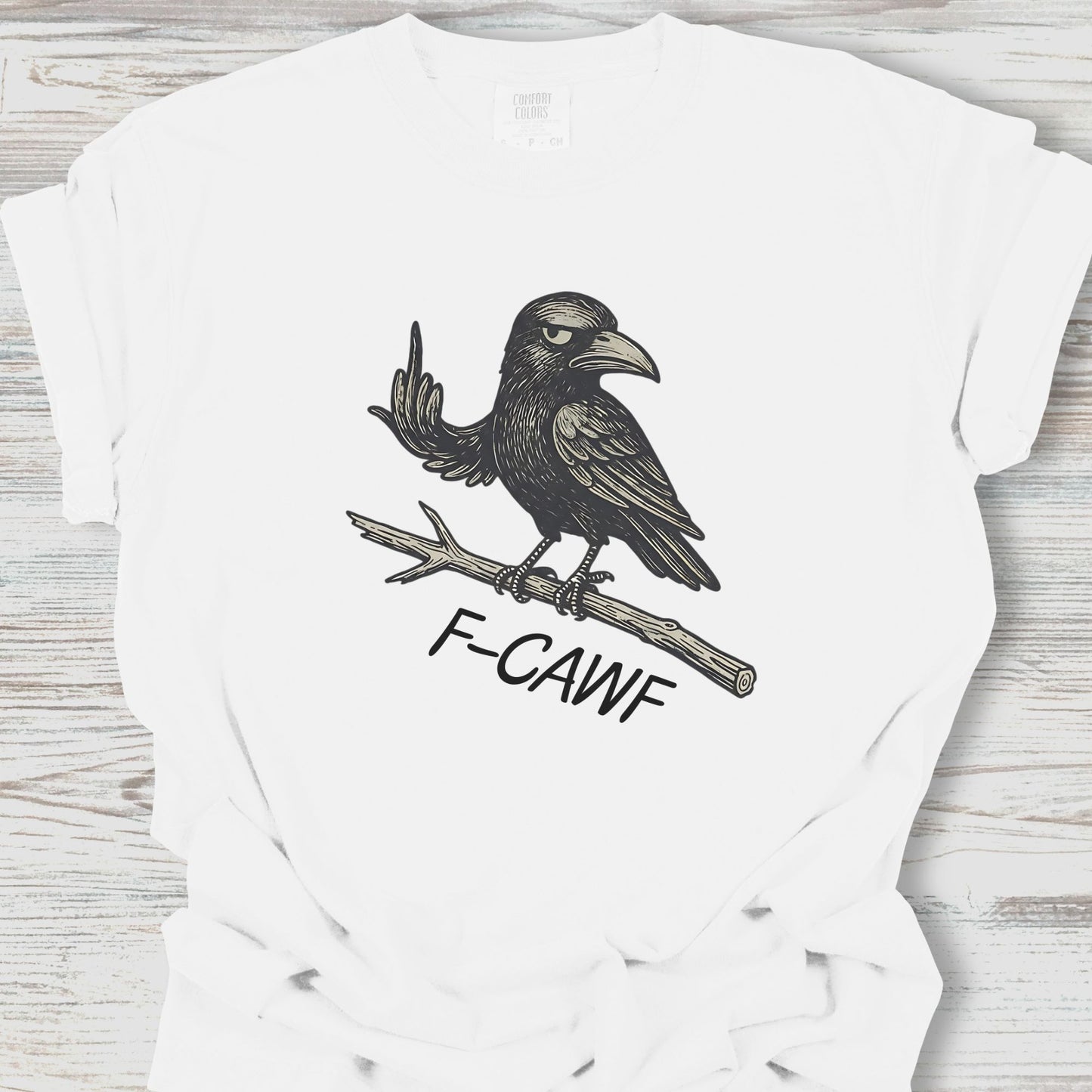 White t-shirt with a black crow illustration and 'F-CAWF' text on a wooden surface
