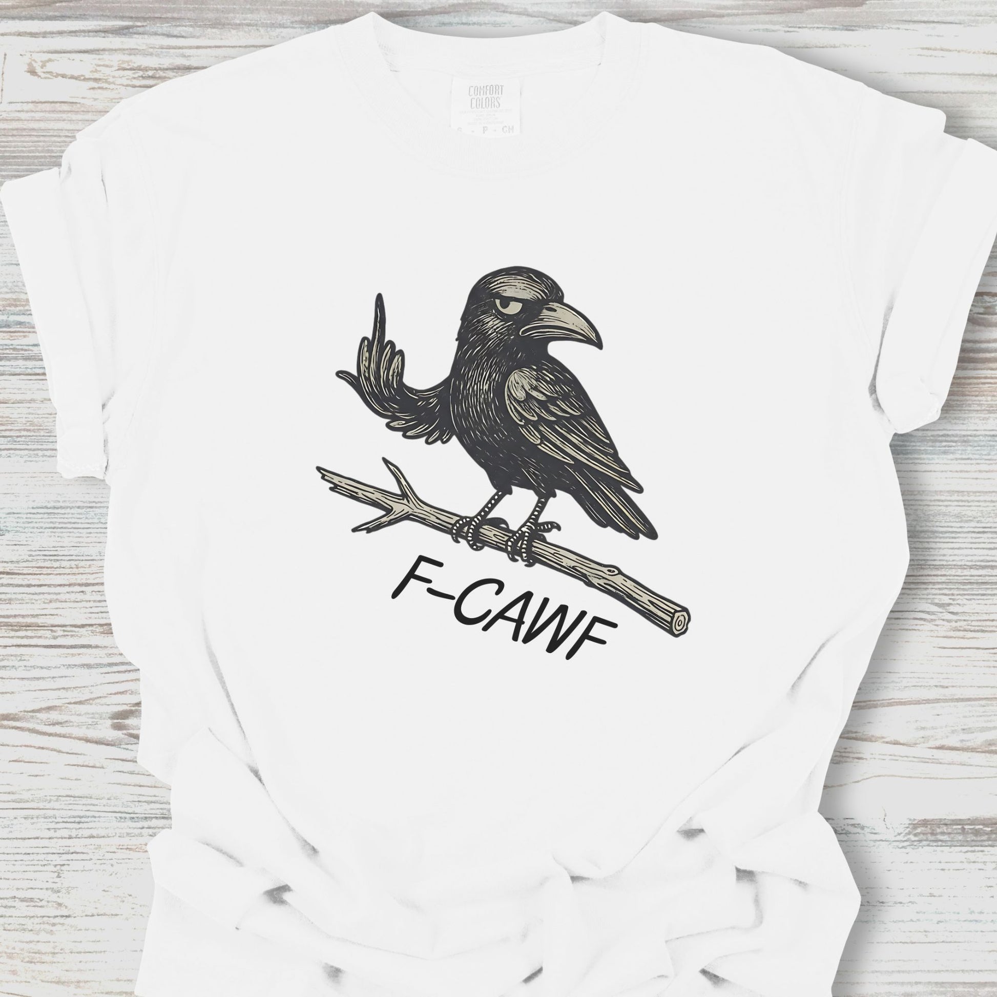 White t-shirt with a black crow illustration and 'F-CAWF' text on a wooden surface