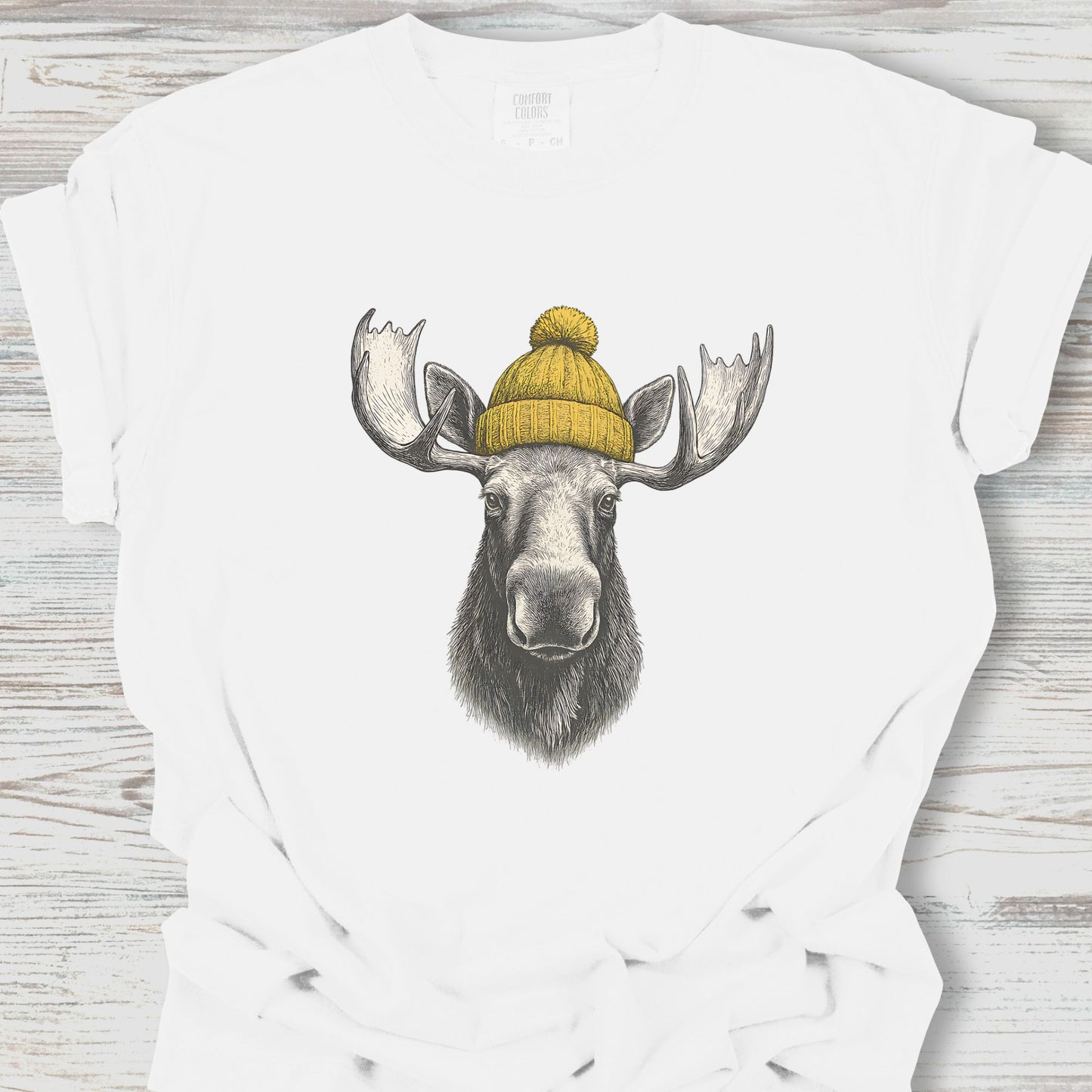 White t-shirt with a moose wearing a yellow beanie design on a wooden surface