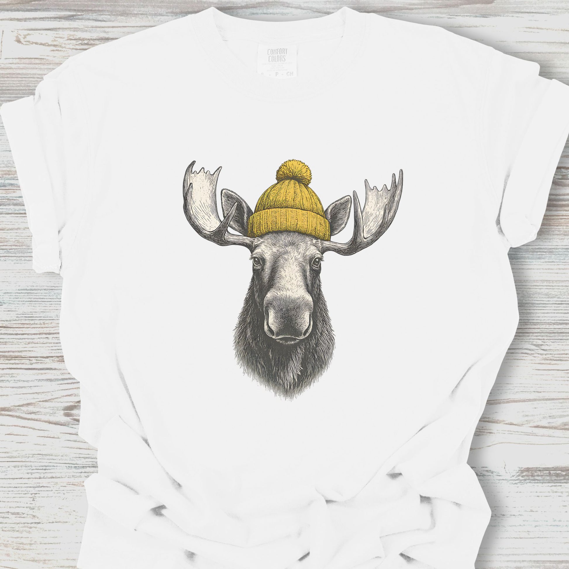 White t-shirt with a moose wearing a yellow beanie design on a wooden surface