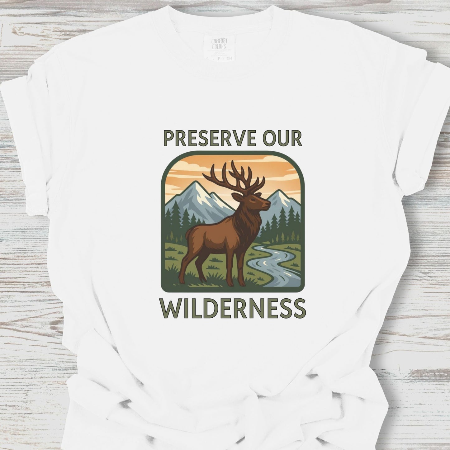 White t-shirt with a graphic of a deer and mountains, and the text 'Preserve Our Wilderness' on a wooden surface.