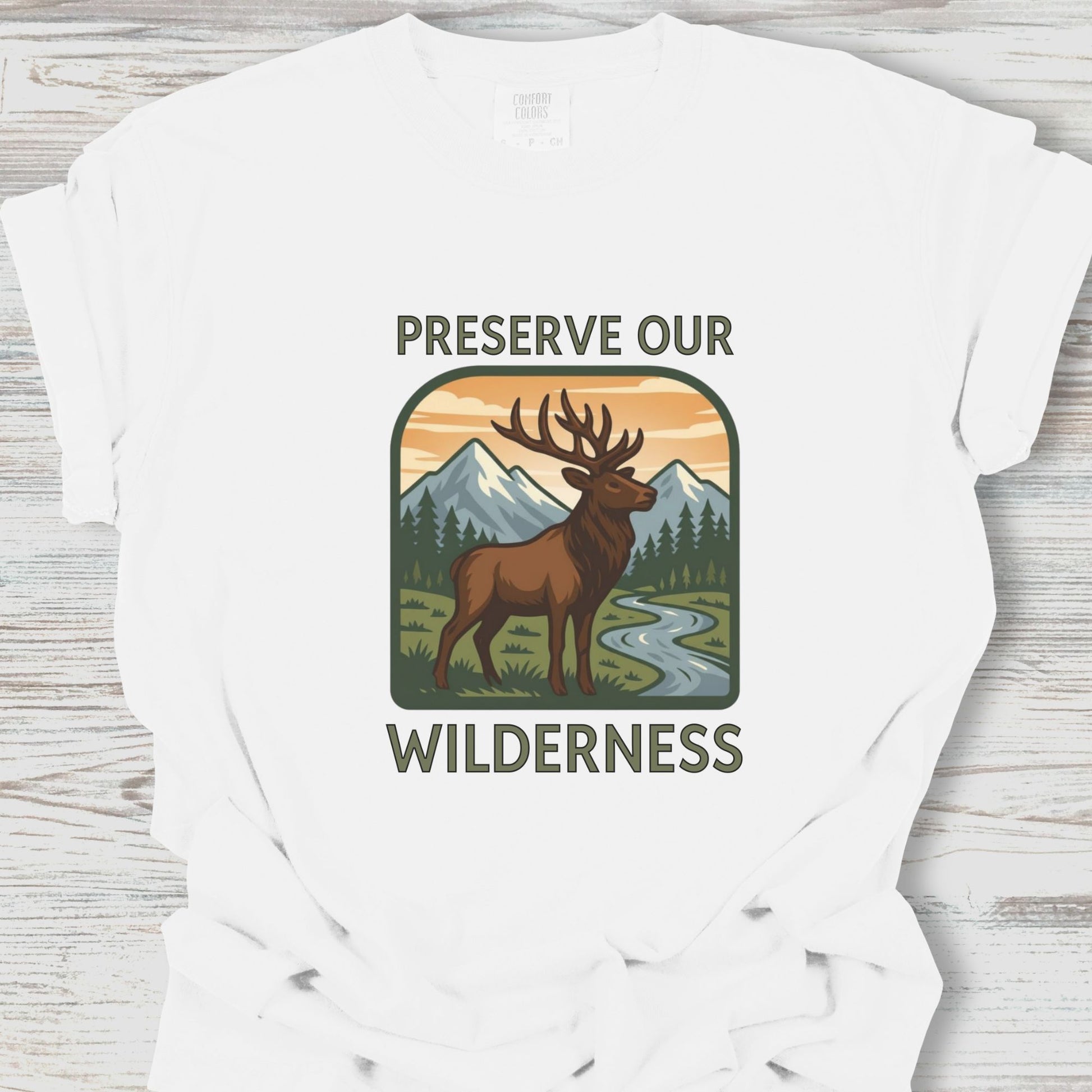 White t-shirt with a graphic of a deer and mountains, and the text 'Preserve Our Wilderness' on a wooden surface.