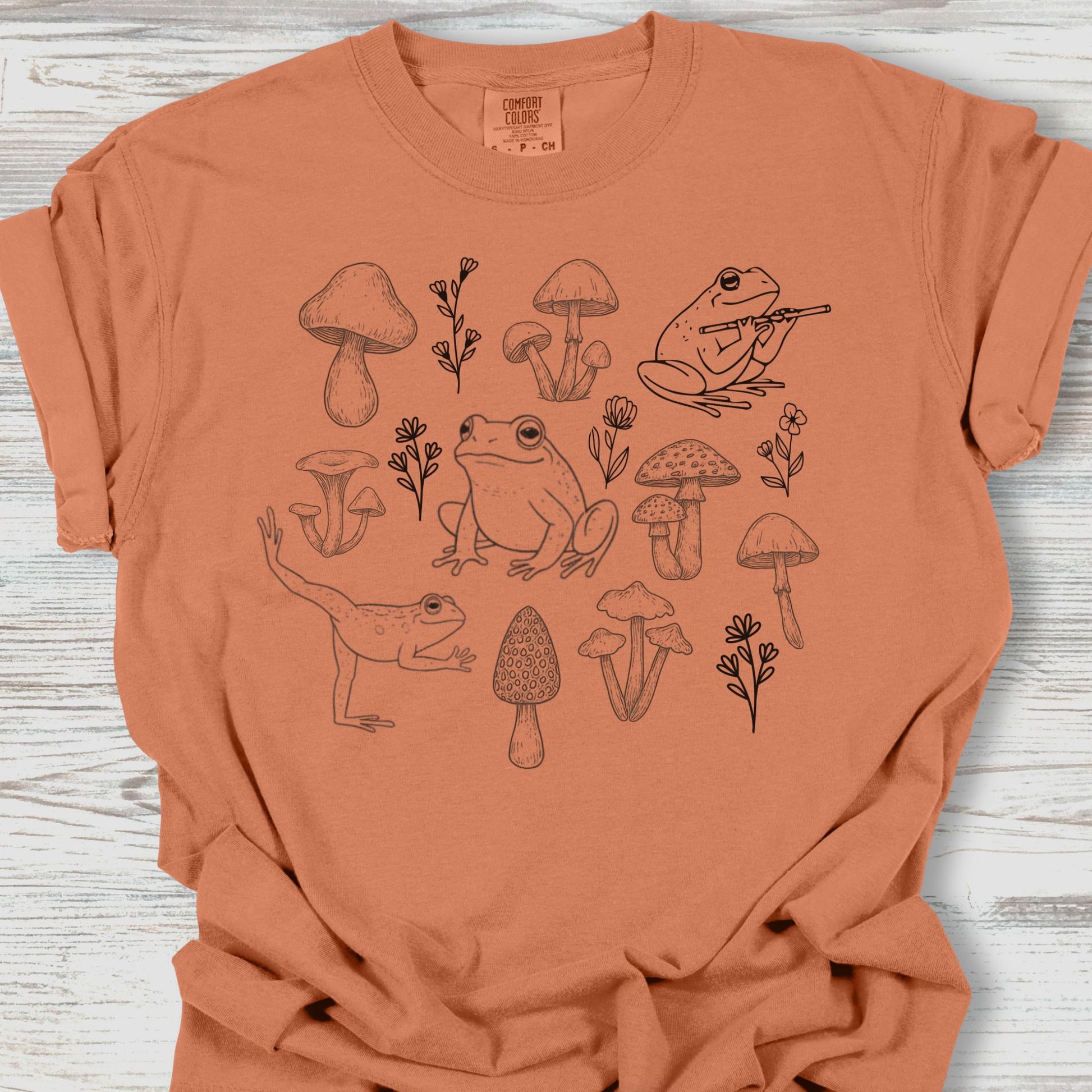 Brown t-shirt with frog and mushroom design on a wooden surface